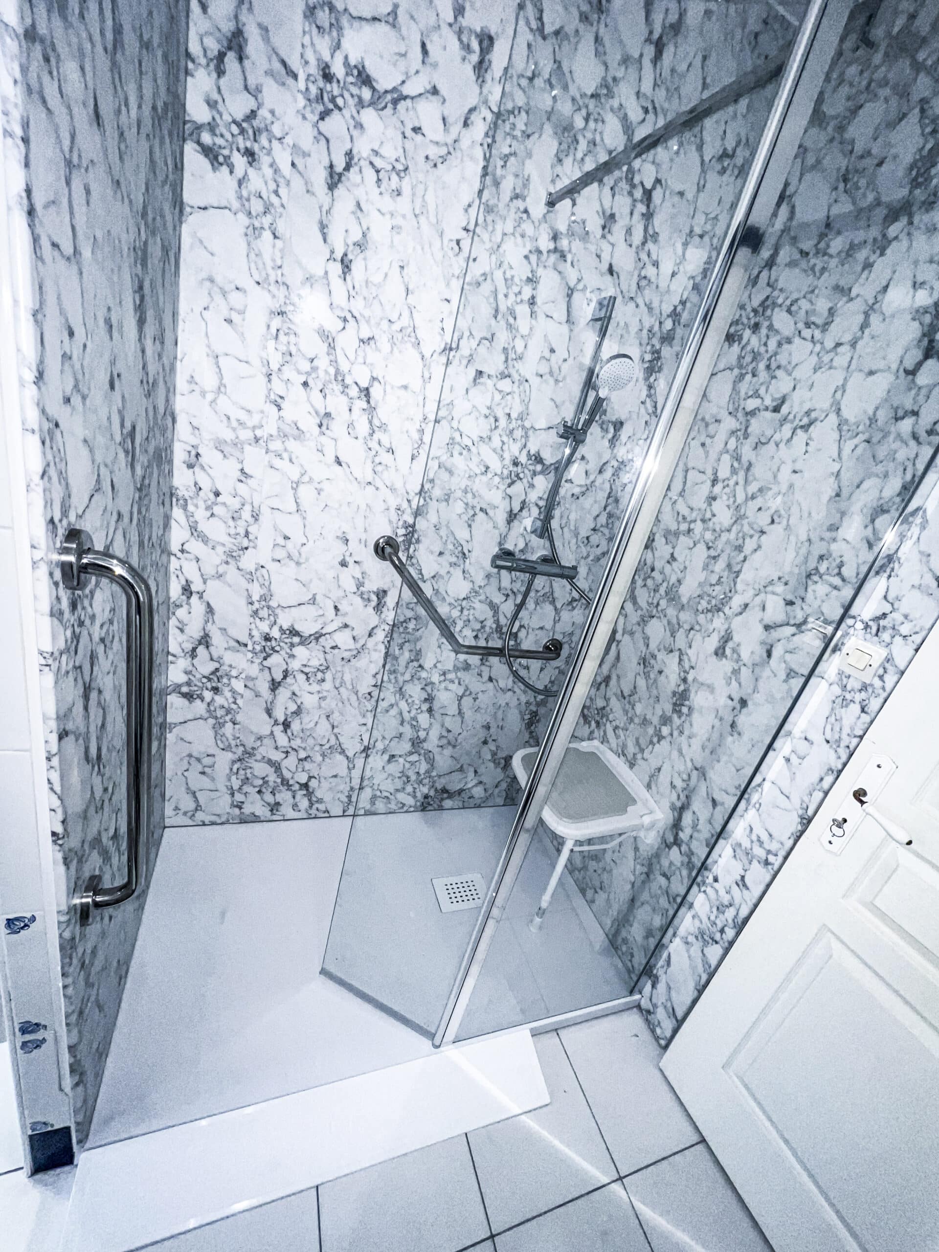 Douche à l'italienne moderne avec murs en marbre blanc et gris, paroi vitrée, pommeau, barres d'appui et siège pliable. Sol clair et porte blanche.