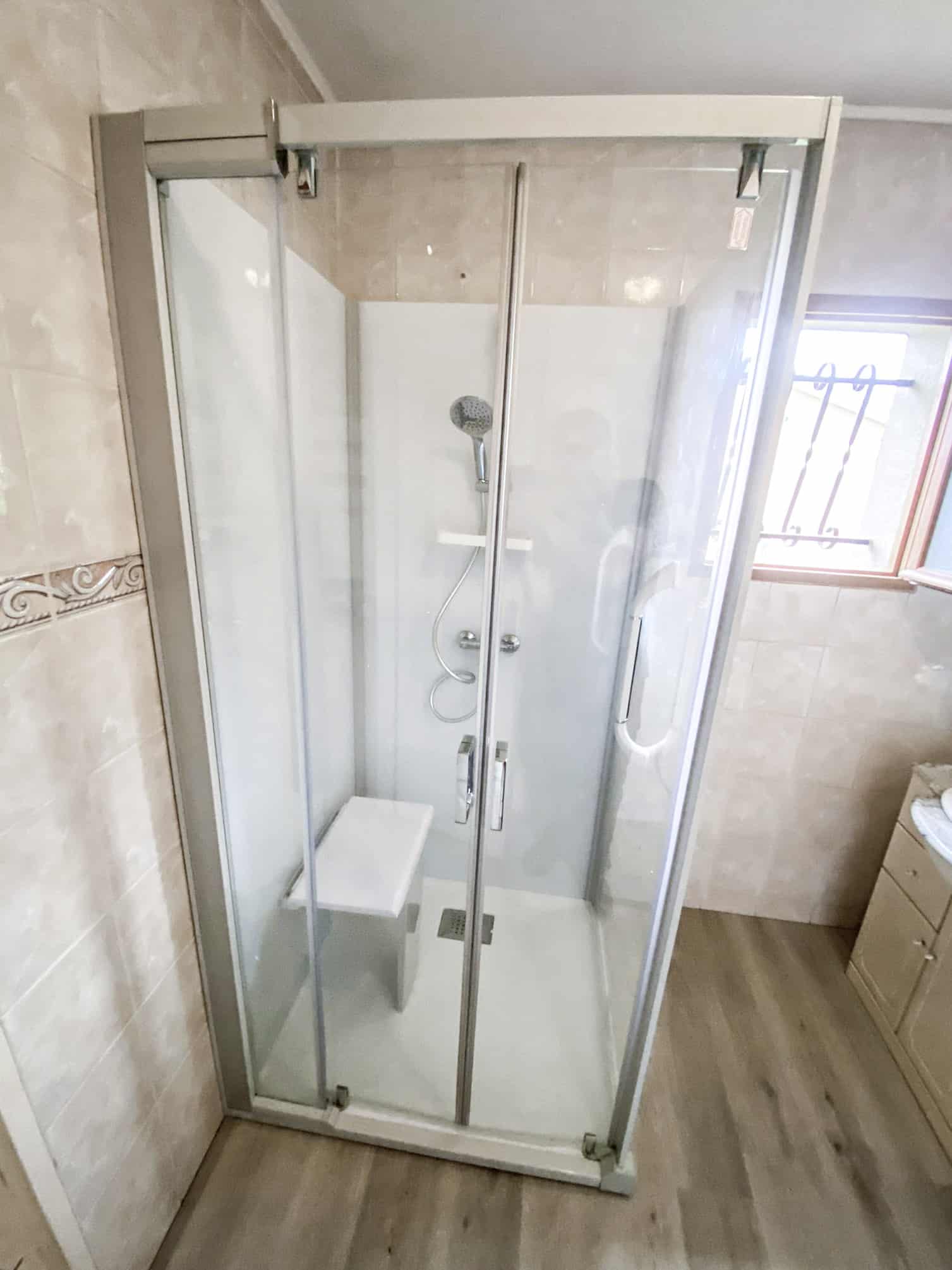 Cabine de douche moderne en verre avec siège intégré et pommeau, dans une salle de bain carrelée beige et sol effet bois.