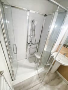Cabine de douche moderne avec parois en verre, pommeau, barres d'appui et siège intégré. Un lavabo sur pied est visible à droite.