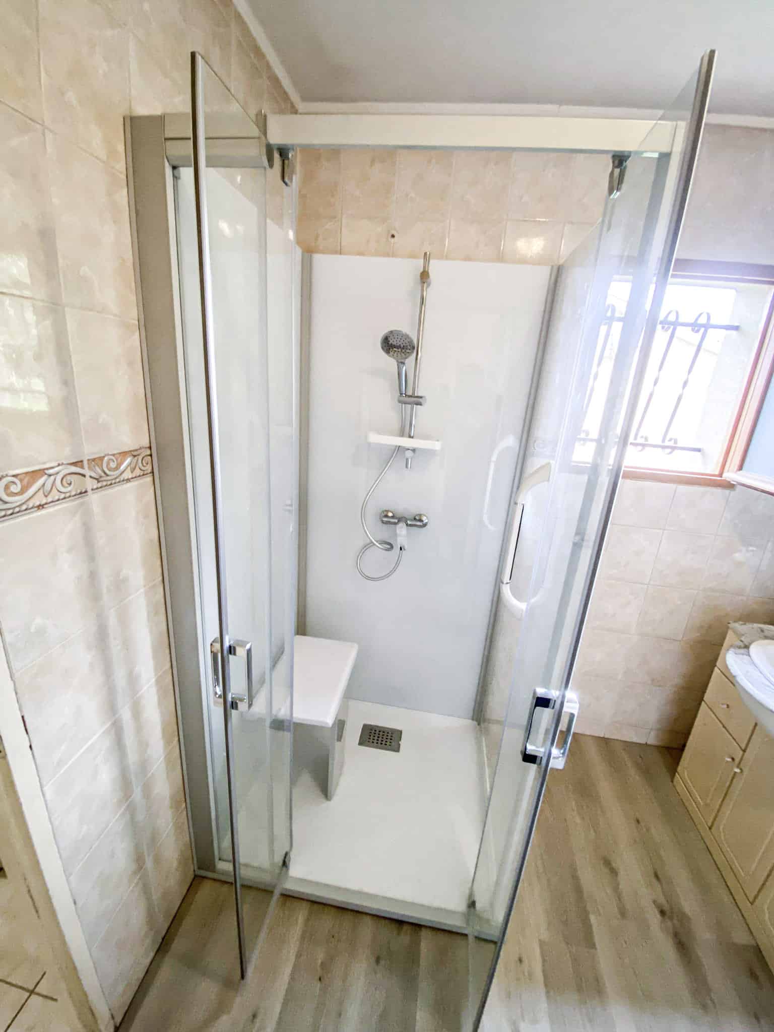 Douche moderne avec parois vitrées, siège et receveur blancs, pommeau chrome. Carrelage mural beige et sol imitation bois clair.