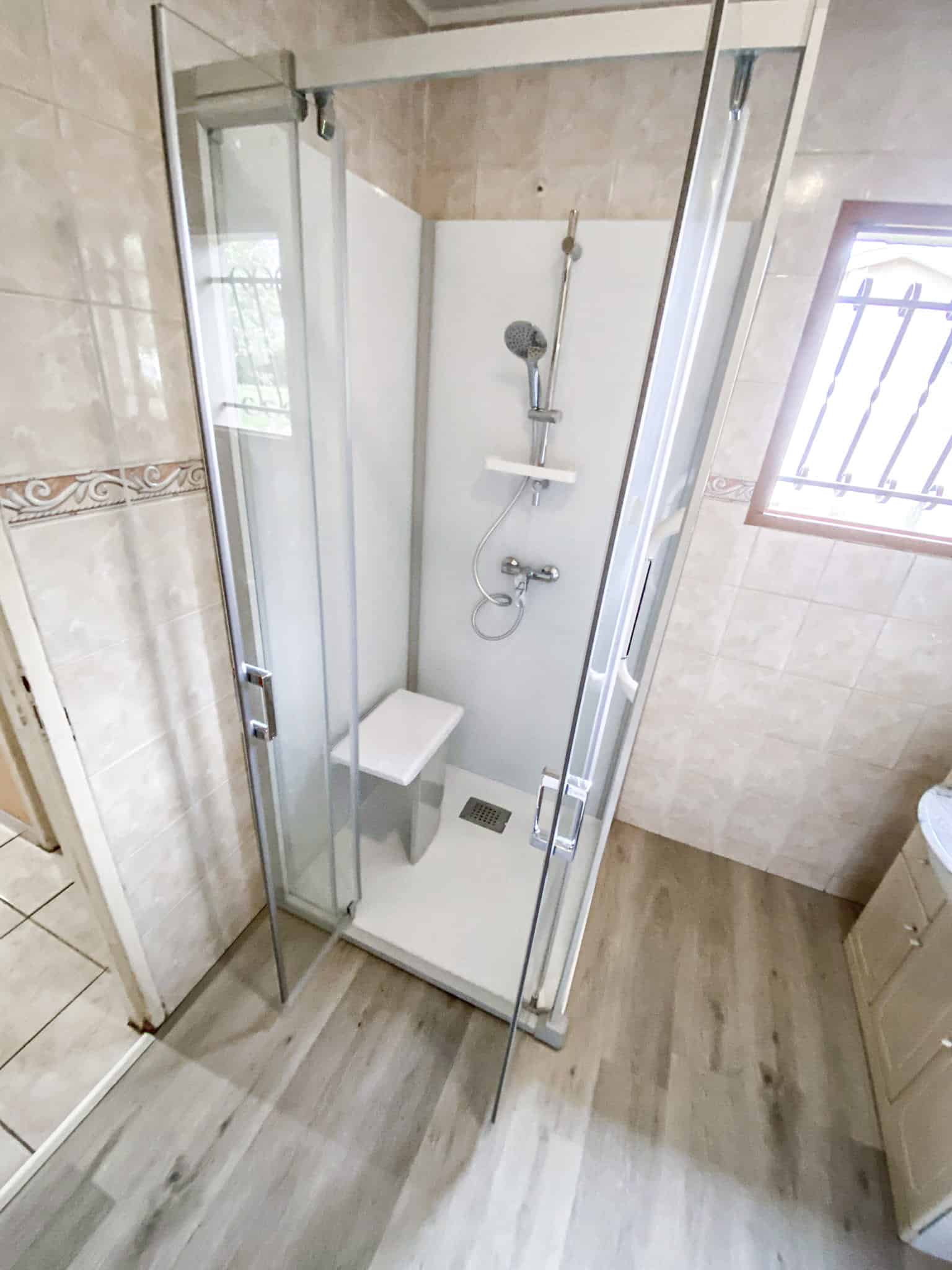 Cabine de douche moderne en verre avec siège intégré, pommeau et mitigeur. SDB avec carrelage beige, sol effet bois et fenêtre à barreaux.