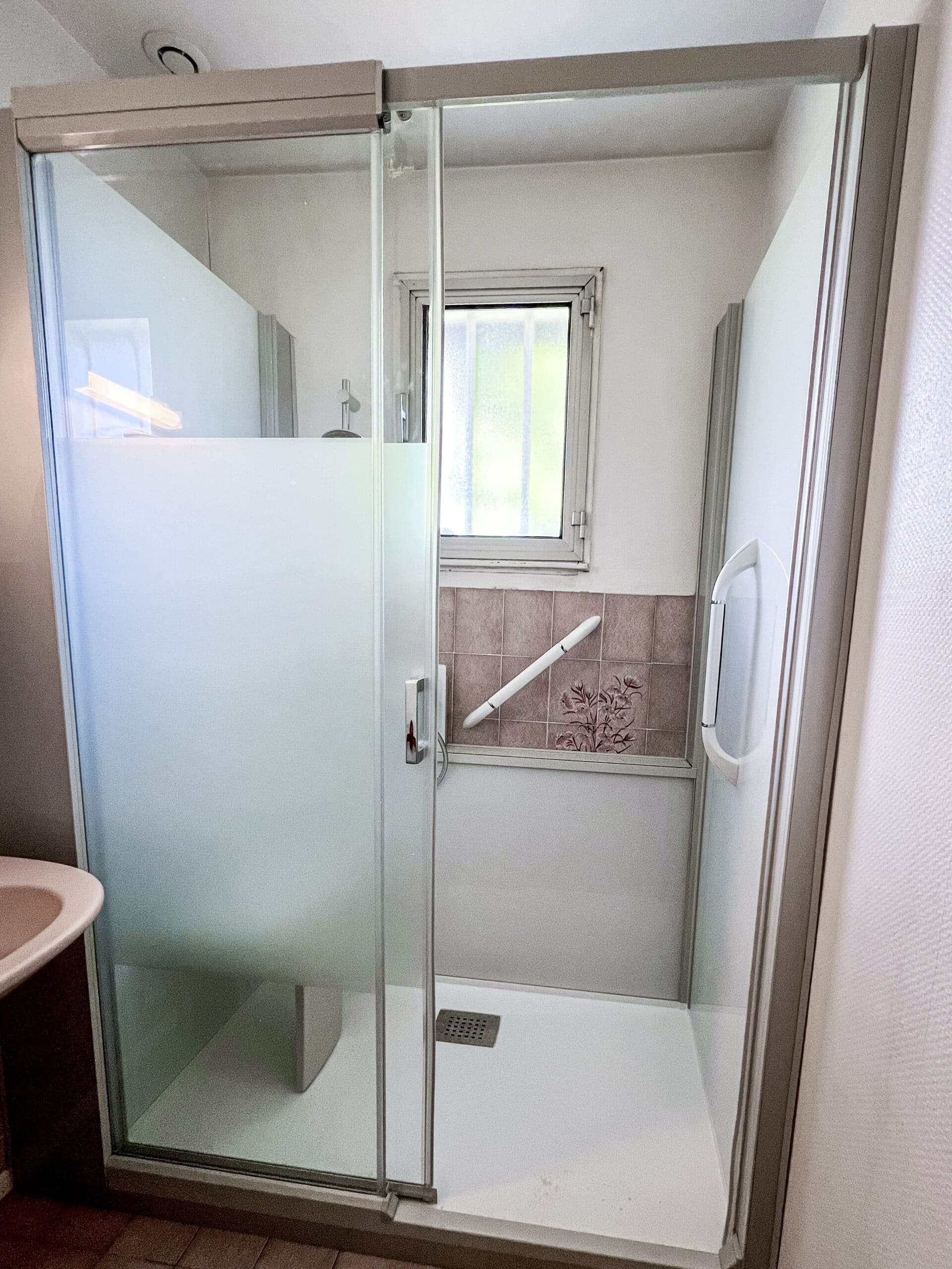 Cabine de douche moderne avec parois en verre dépoli, porte coulissante, receveur blanc, barre d'appui et carrelage rose floral.