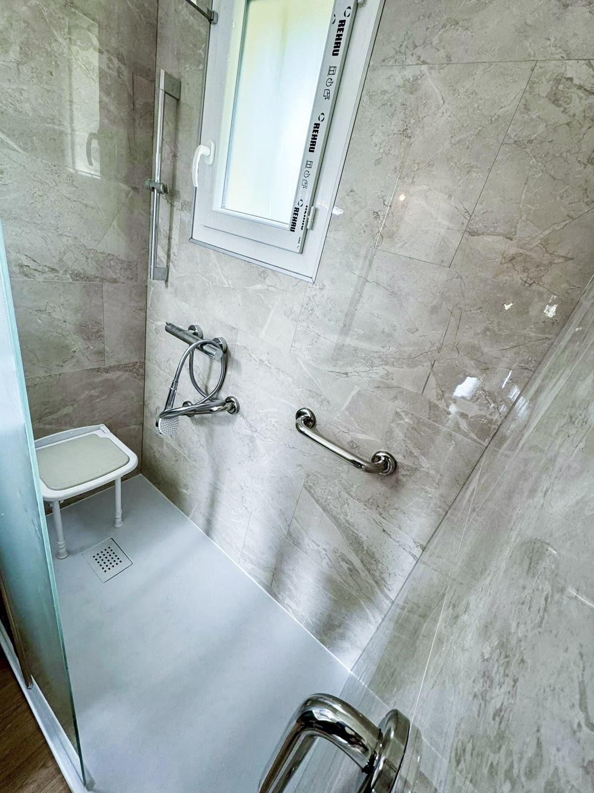 Douche moderne accessible avec murs carrelés gris marbré, siège de douche, barre d'appui chromée et fenêtre REHAU. Bac à douche clair et drain au sol.