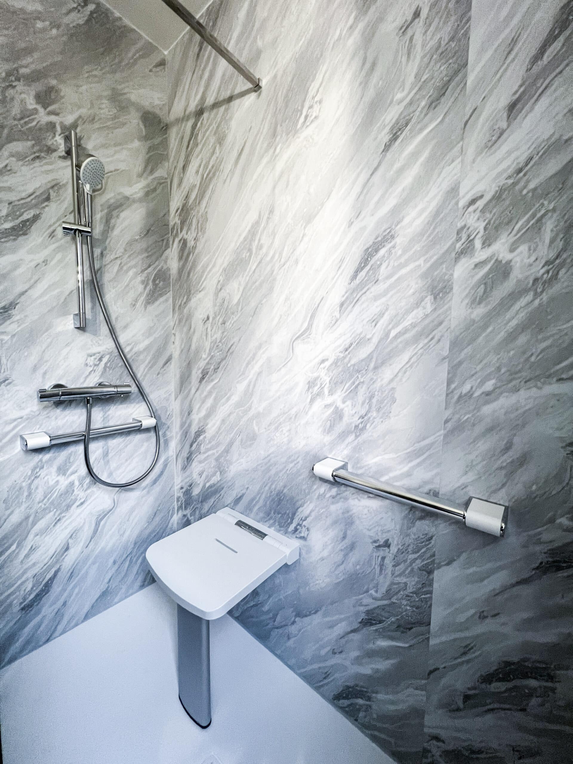 Douche PMR moderne avec murs imitation marbre gris. Siège rabattable blanc, barre d'appui chromée et pommeau de douche mural. Sol antidérapant.