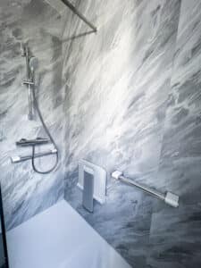 Douche accessible moderne avec murs aspect marbre gris, pommeau réglable, mitigeur, siège pliant et barre d'appui chromée.