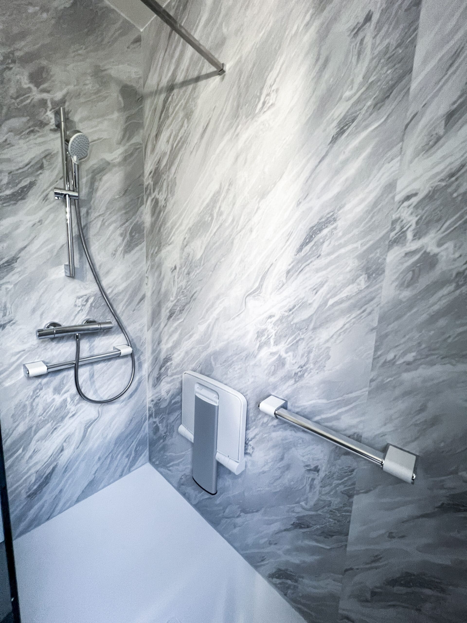 Douche accessible moderne avec murs aspect marbre gris, pommeau réglable, mitigeur, siège pliant et barre d'appui chromée.