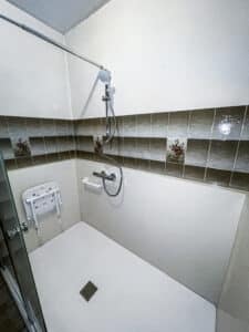 Douche moderne et accessible avec carrelage marron floral, siège rabattable, barre d'appui et pommeau de douche mural. Sol et murs blancs.
