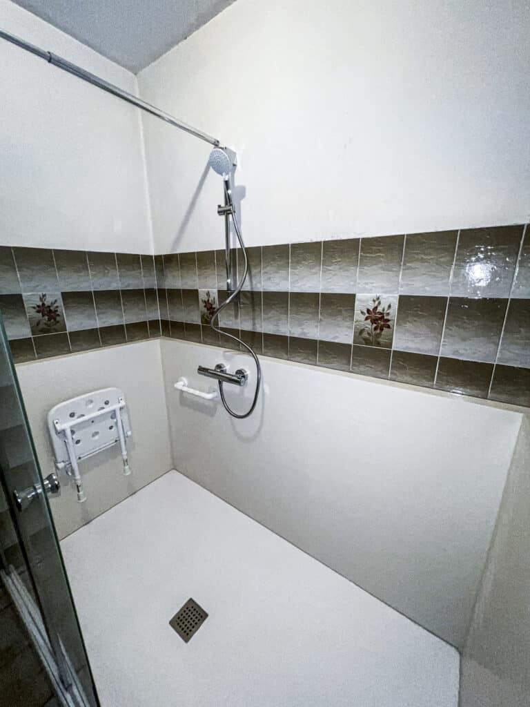 Douche moderne et accessible avec carrelage marron floral, siège rabattable, barre d'appui et pommeau de douche mural. Sol et murs blancs.