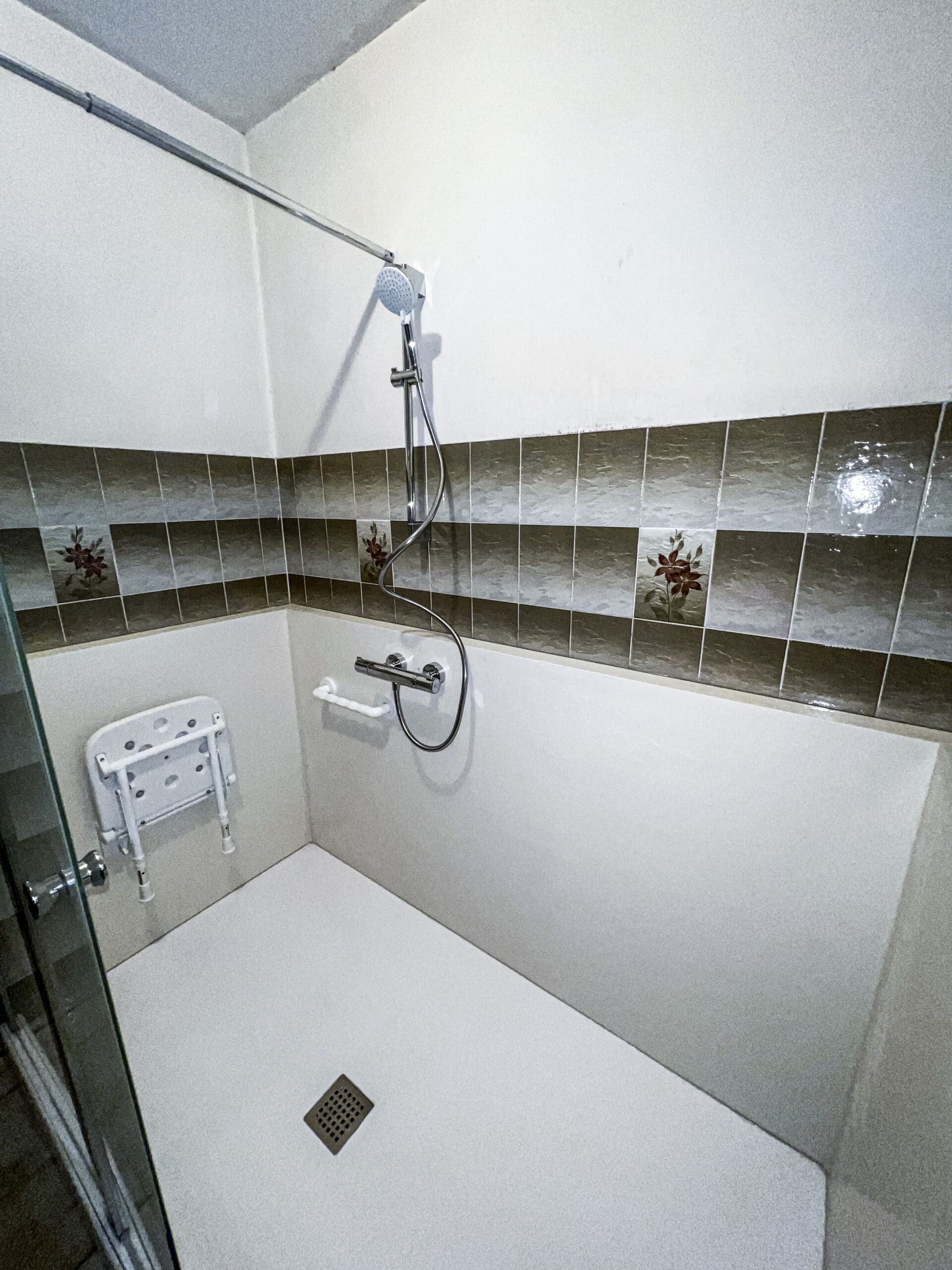 Douche moderne et accessible avec carrelage marron floral, siège rabattable, barre d'appui et pommeau de douche mural. Sol et murs blancs.