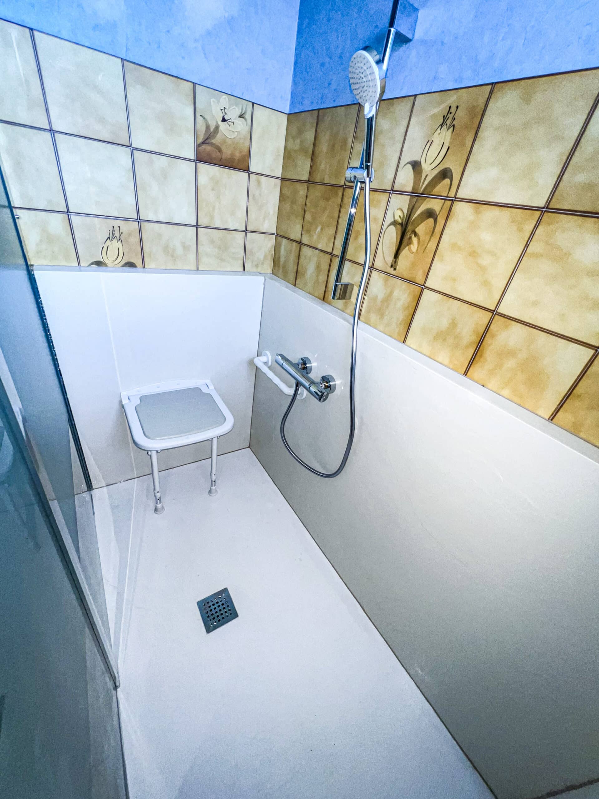 Douche moderne accessible avec murs carrelés motifs floraux, siège rabattable, pommeau de douche et receveur blanc. Mur supérieur bleu.
