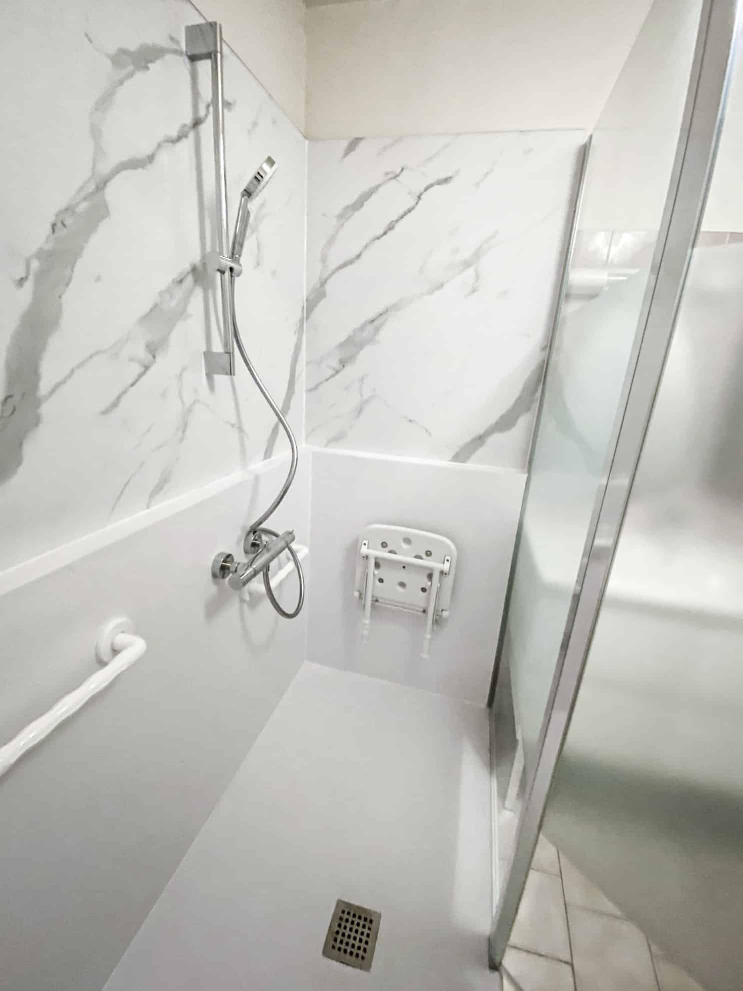 Douche moderne et accessible avec carrelage marbré, barre d'appui blanche et siège pliable. Finitions chromées.