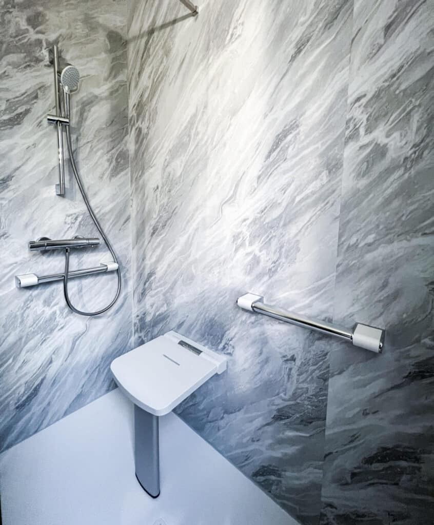 Douche adaptée avec parois effet marbre gris, siège de douche rabattable blanc et barre d'appui chromée.