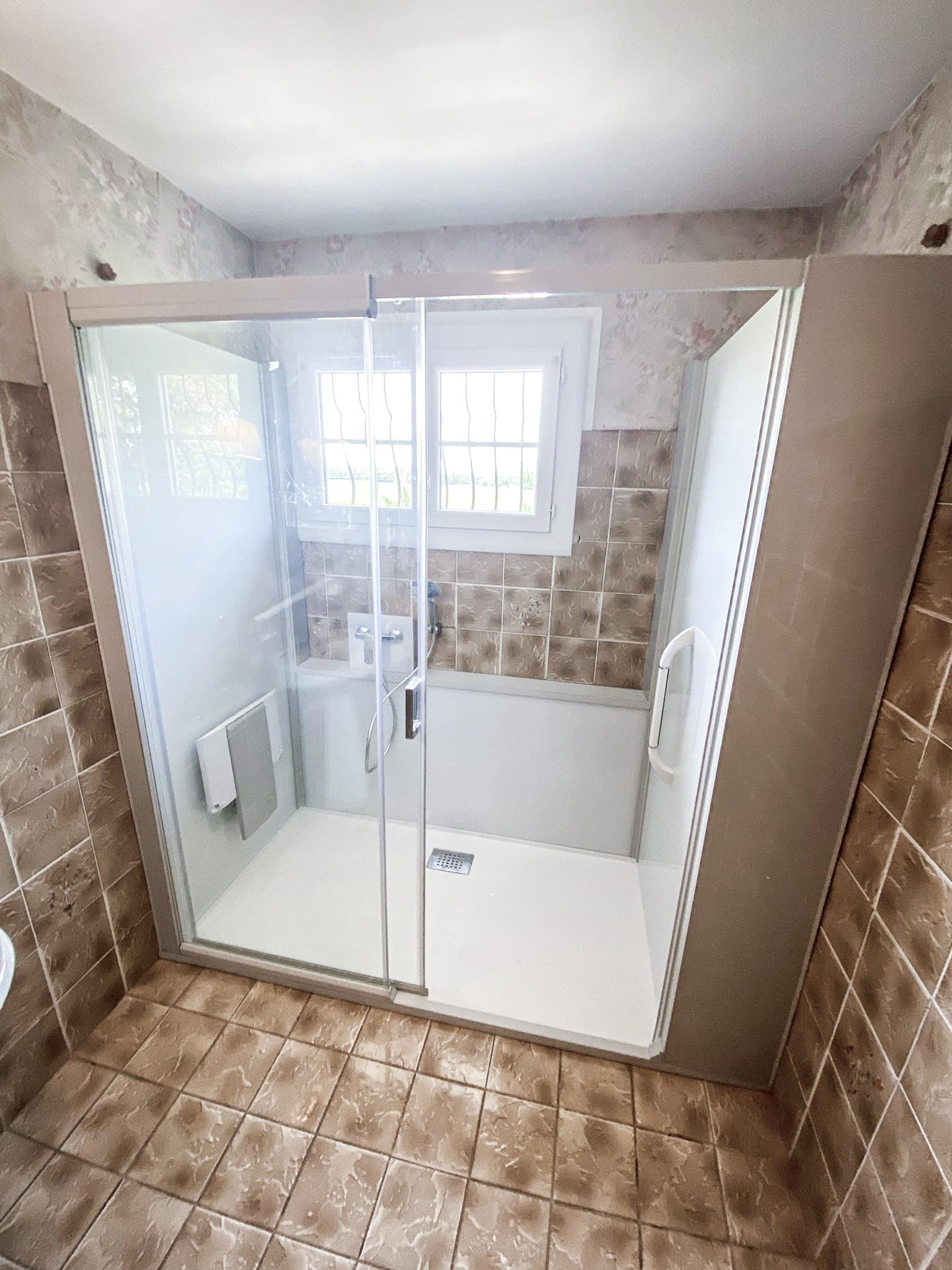 Douche d'angle moderne avec portes en verre transparentes, receveur blanc et murs partiellement carrelés bruns. Fenêtre claire visible à l'intérieur.