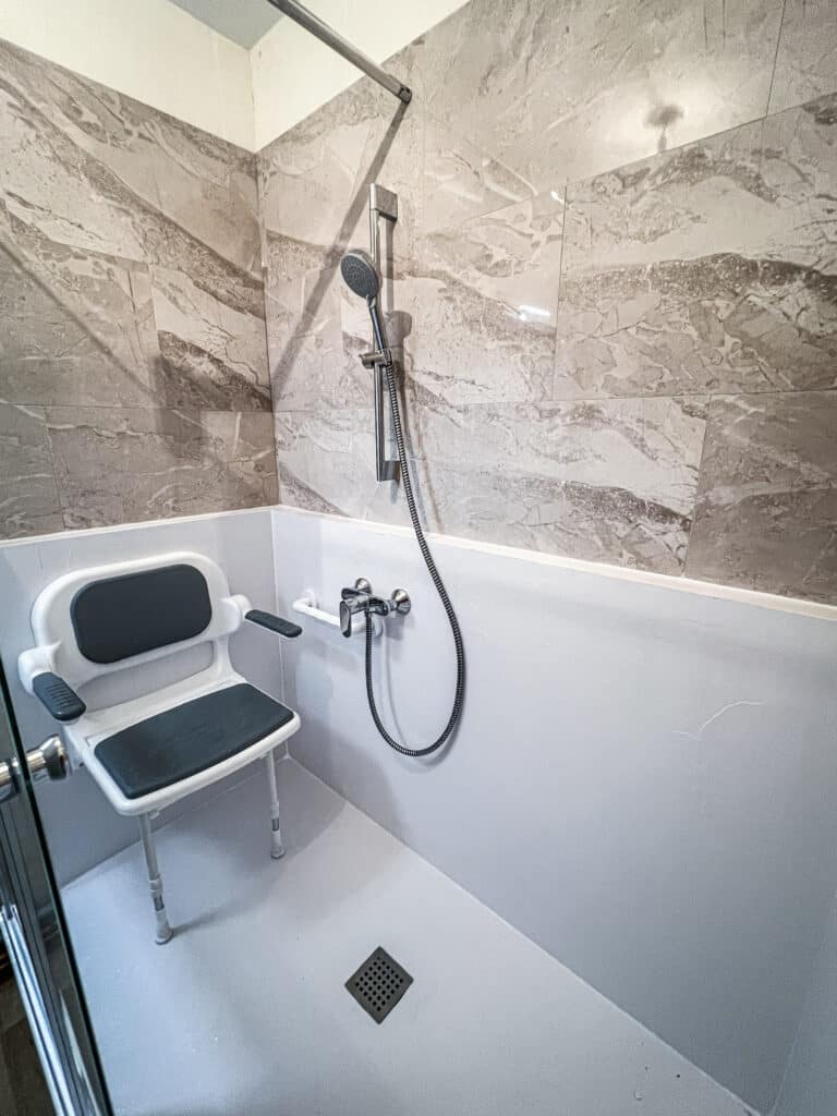 Douche à l'italienne moderne et accessible avec carrelage effet marbre, siège de douche mural, barre d'appui et pommeau à main.