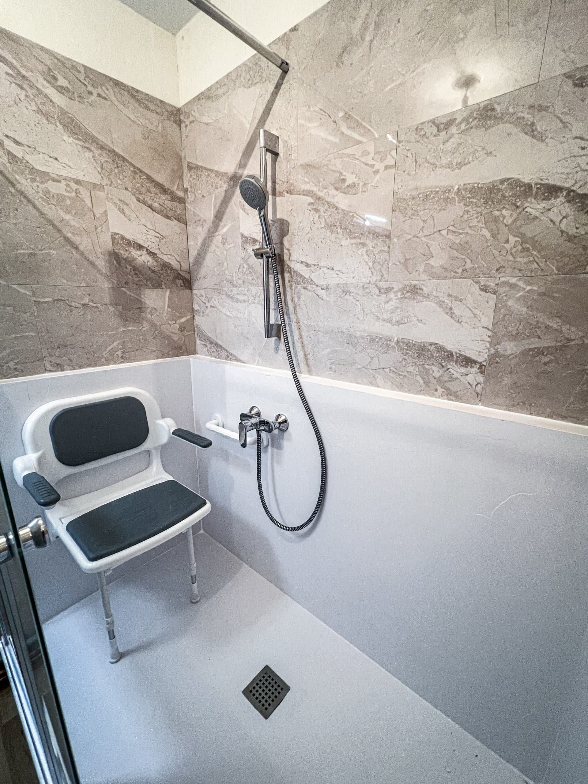 Douche à l'italienne moderne et accessible avec carrelage effet marbre, siège de douche mural, barre d'appui et pommeau à main.