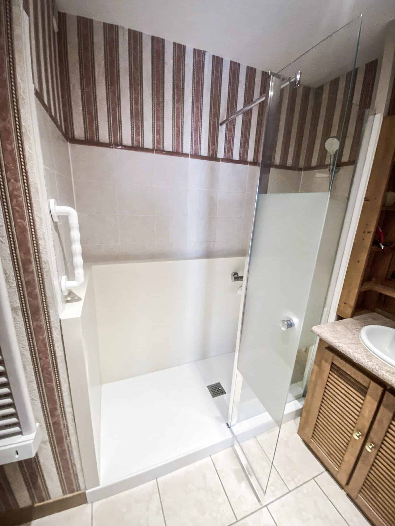 Douche à l'italienne moderne et accessible avec barre d'appui blanche. Murs carrelés, papier peint rayé, et meuble vasque en bois.