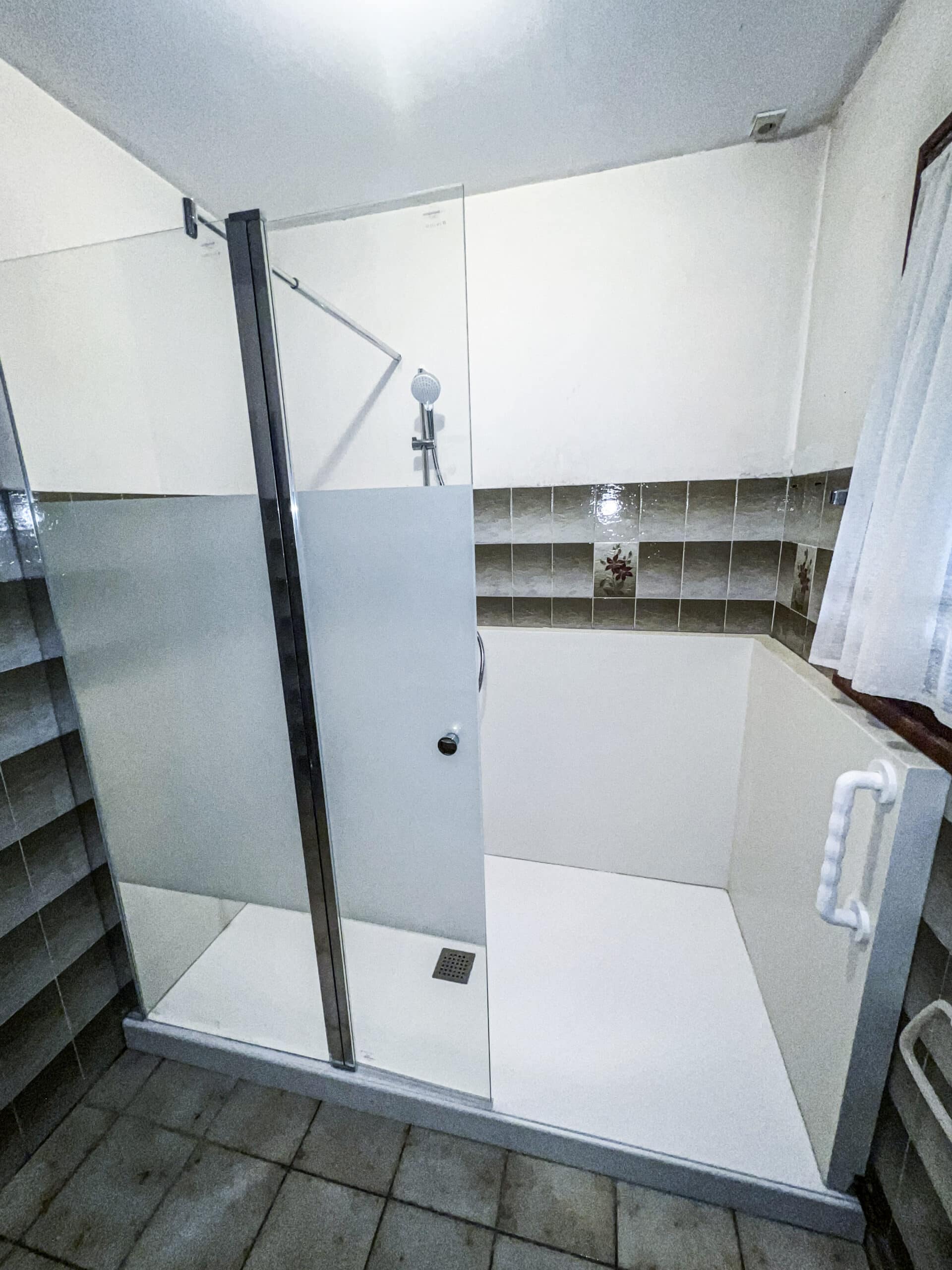 Douche à l'italienne moderne avec paroi vitrée, receveur blanc, barre d'appui et carrelage mural marron dans une salle de bain lumineuse.