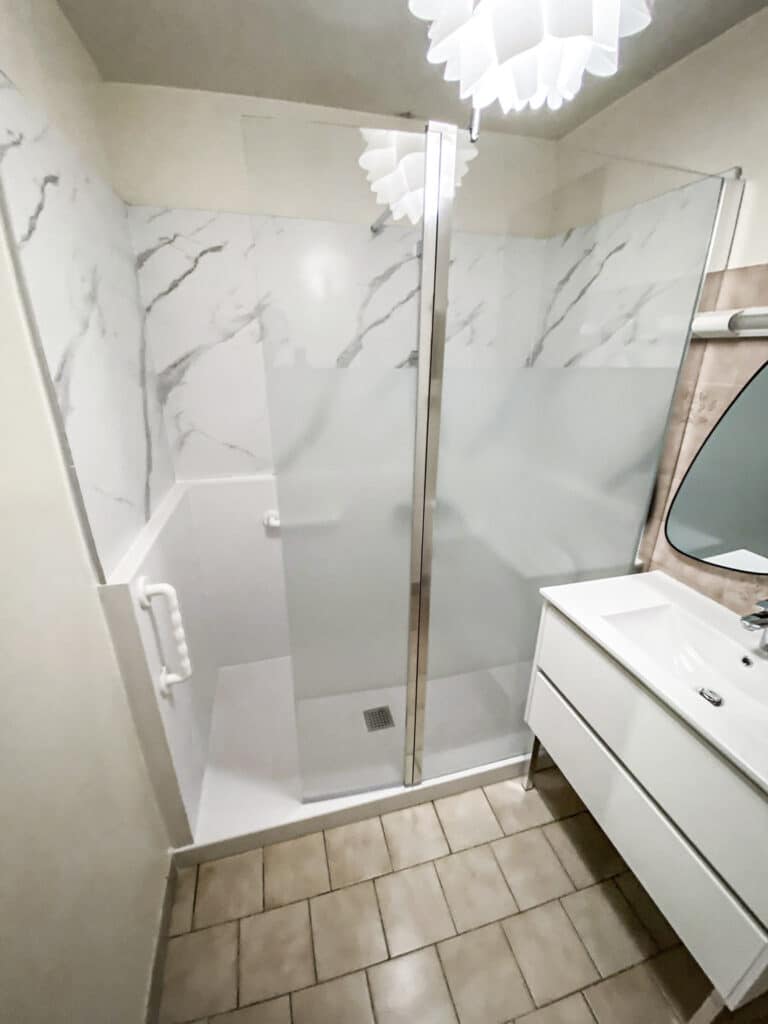 Douche à l'italienne moderne et salle de bain fonctionnelle Salle de bain moderne avec douche à l'italienne carrelée aspect marbre, paroi vitrée, barre d'appui. Meuble-vasque blanc et miroir.