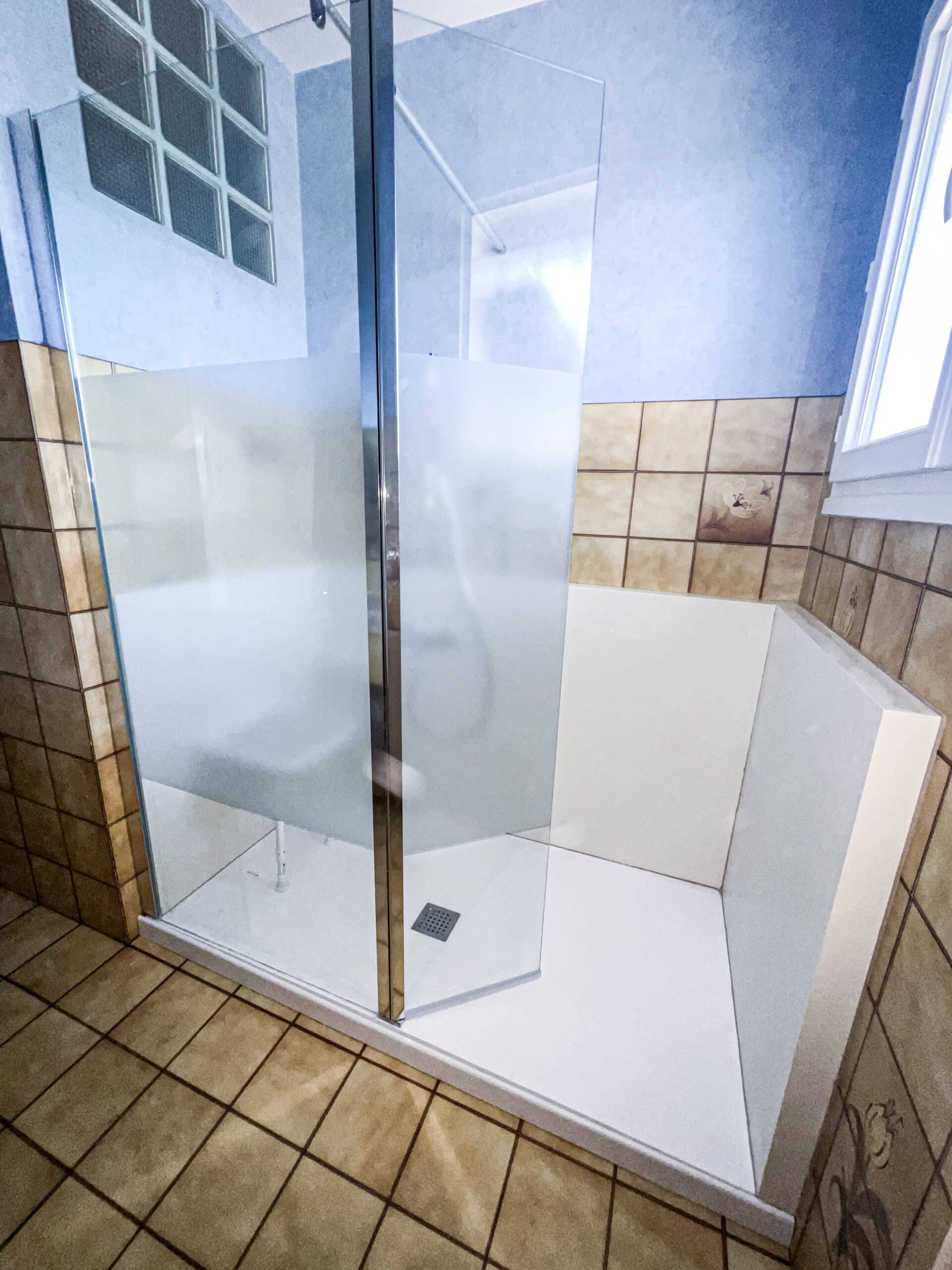 Salle de bain moderne avec douche à l'italienne en verre. Murs carrelés beige, partie haute bleue. Fenêtre et blocs de verre.