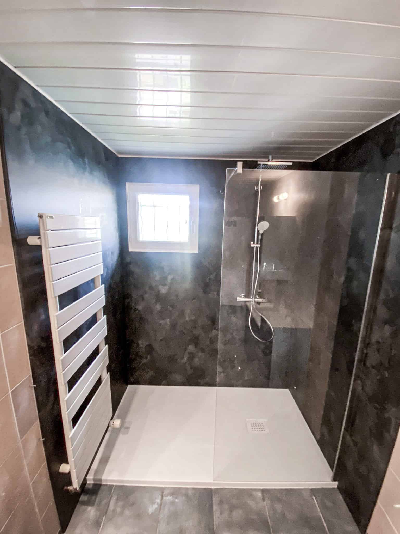 Salle de bain sombre avec douche à l'italienne vitrée, receveur blanc, radiateur sèche-serviettes et murs effet béton noir.