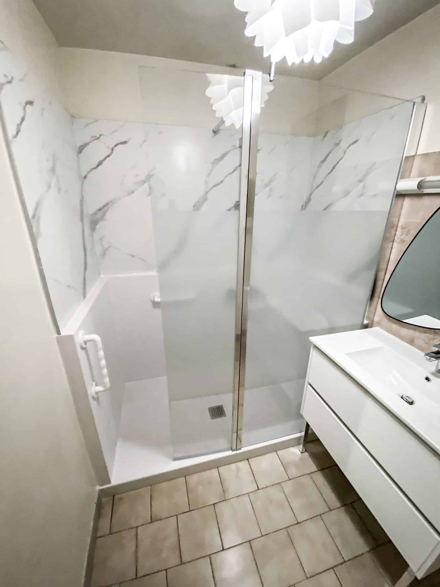 Salle de bain moderne avec douche à l'italienne carrelée aspect marbre, paroi vitrée, barre d'appui. Meuble-vasque blanc et miroir.