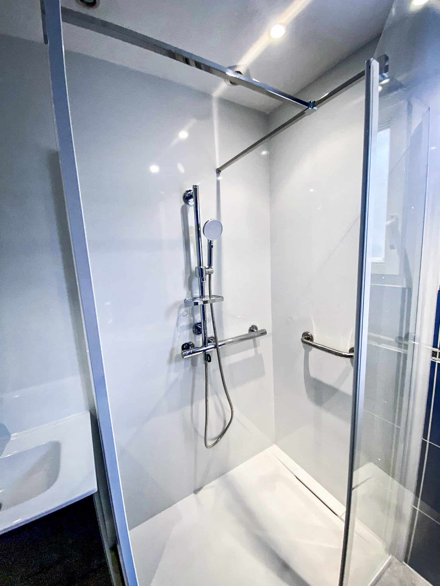 Douche moderne et accessible avec parois blanches, receveur de plain-pied, système chromé et deux barres d'appui. Cabine de douche en verre.
