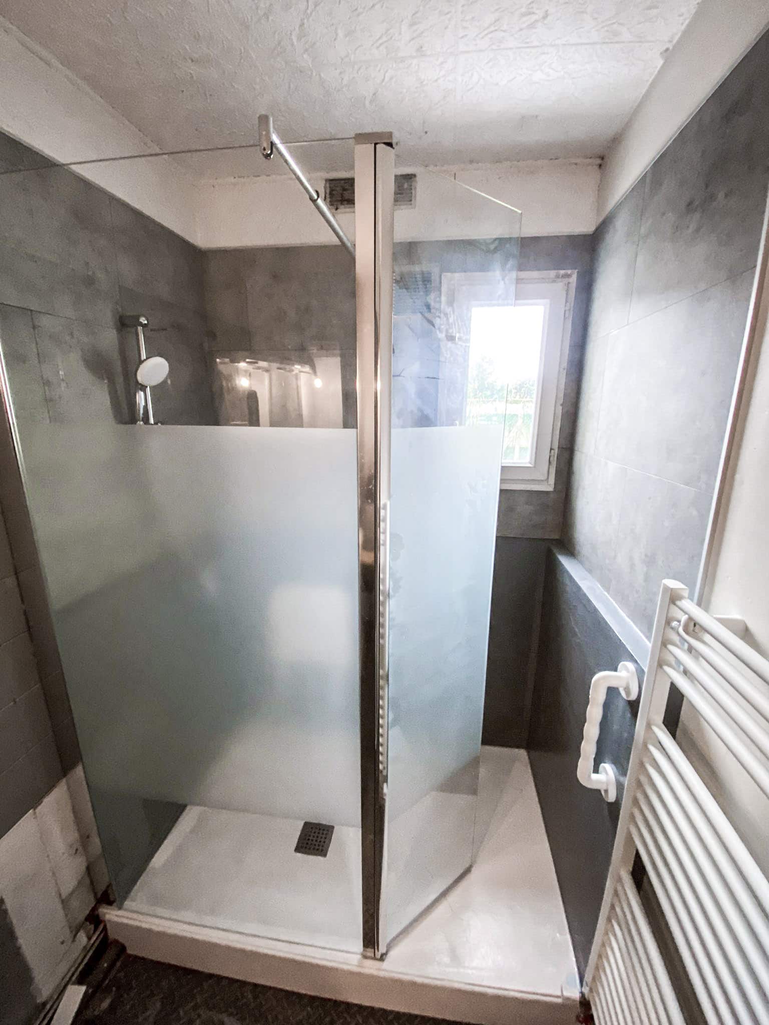 Douche moderne avec paroi vitrée dépolie, carrelage mural gris foncé et receveur blanc. Une fenêtre et un radiateur sèche-serviettes sont visibles.