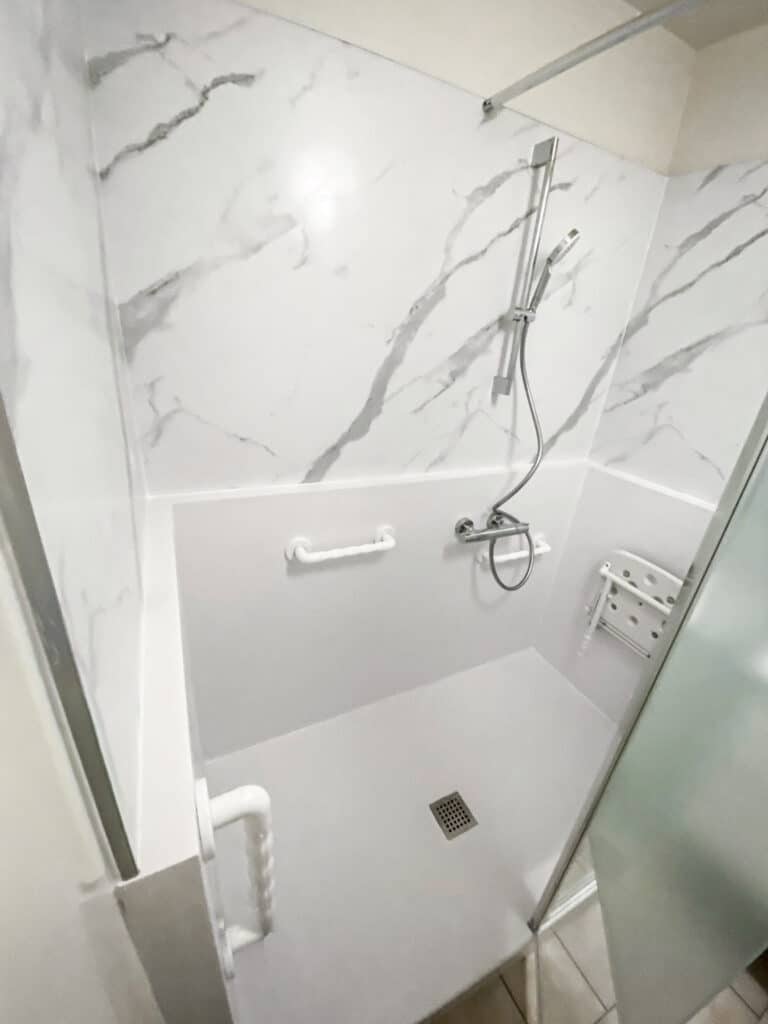 Douche moderne et accessible avec parois effet marbre blanc/gris. Équipée d'une pomme de douche, barre d'appui, siège rabattable blanc et drain au sol.