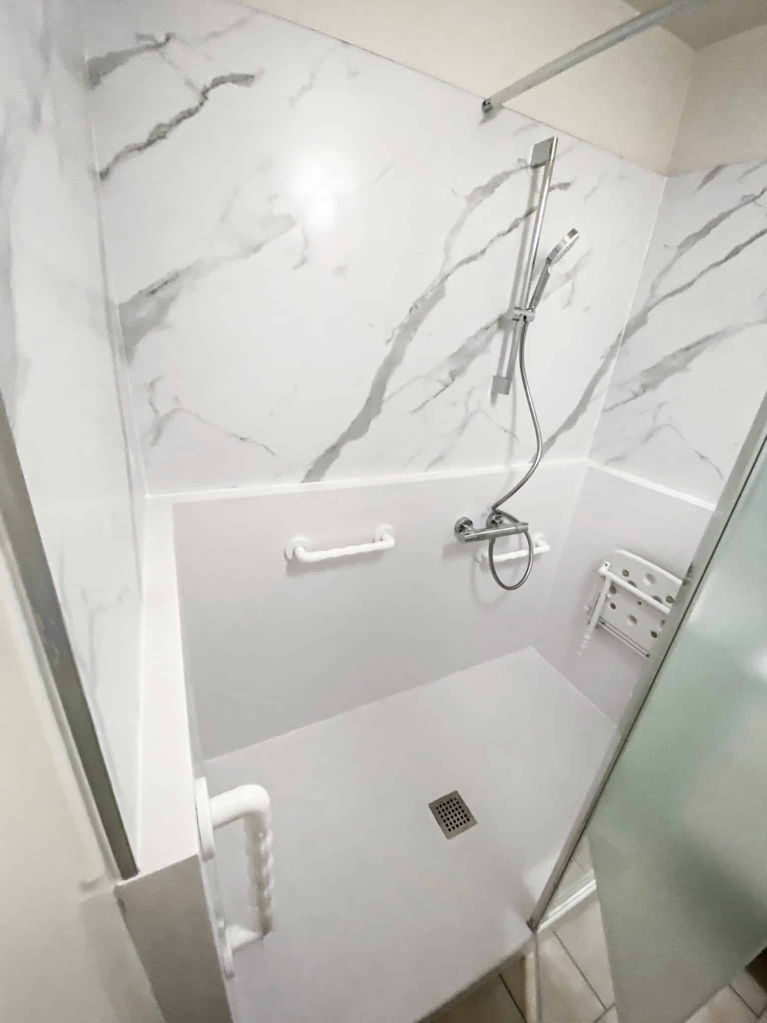 Douche moderne et accessible avec parois effet marbre blanc/gris. Équipée d'une pomme de douche, barre d'appui, siège rabattable blanc et drain au sol.