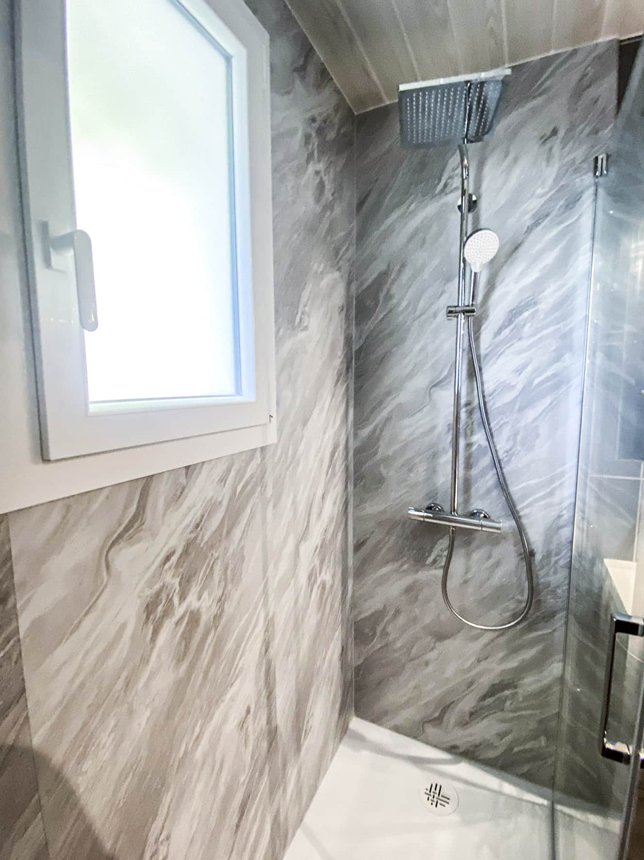 Douche moderne avec parois effet marbre gris, pommeau pluie chromé et douchette, base blanche. Fenêtre lumineuse à gauche.