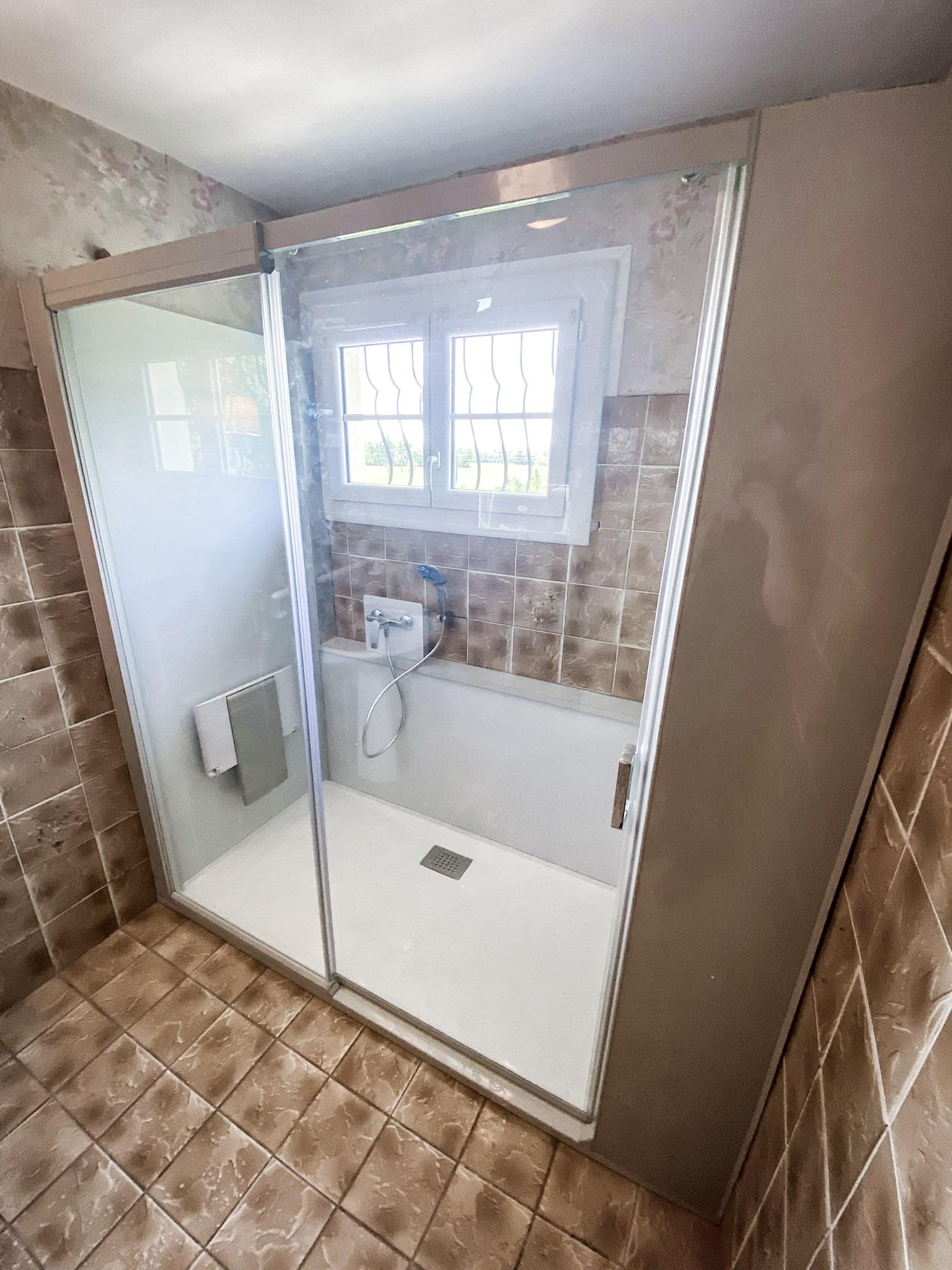 Douche moderne vitrée avec receveur blanc dans une salle de bain carrelée marron. Présence d'une fenêtre et de serviettes.