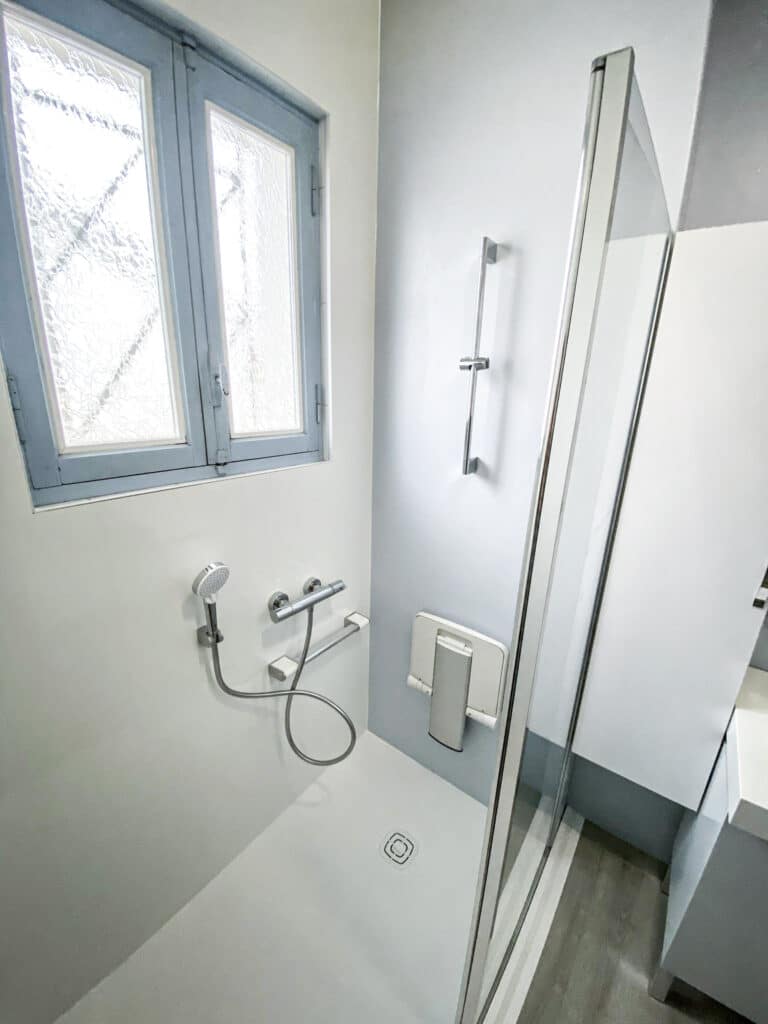 Douche moderne avec siège rabattable et pommeau, fenêtre à vitrage texturé, murs bicolores gris/blanc et cabine en verre.