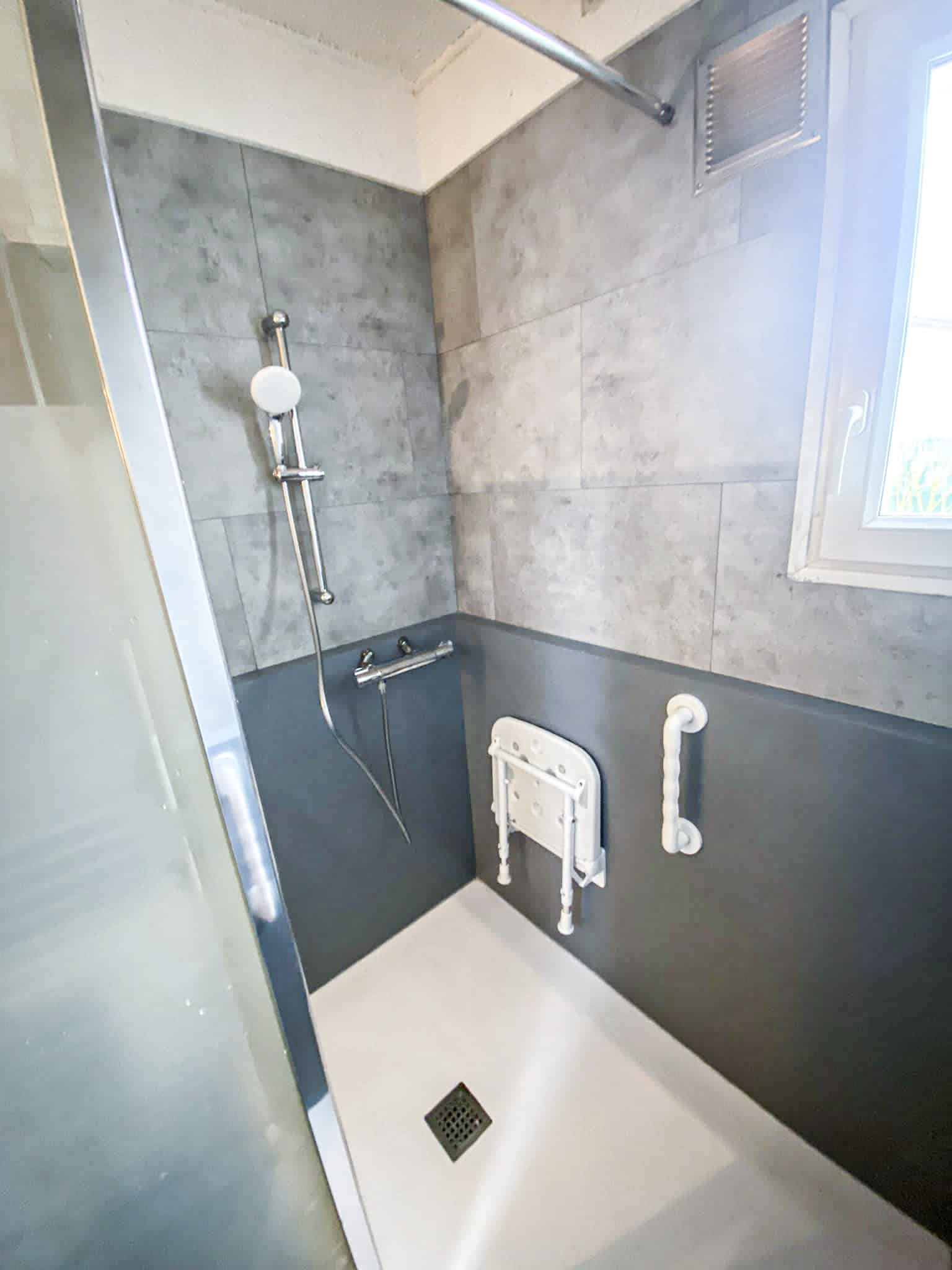 Douche moderne accessible PMR avec carrelage gris, panneaux anthracite, siège pliant, barre d'appui et receveur extra-plat.