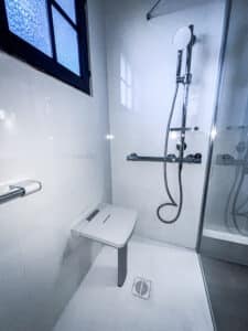 Douche moderne et accessible avec siège rabattable mural, barre d'appui chromée, pommeau réglable et fenêtre à verre bleu texturé. Espace épuré.