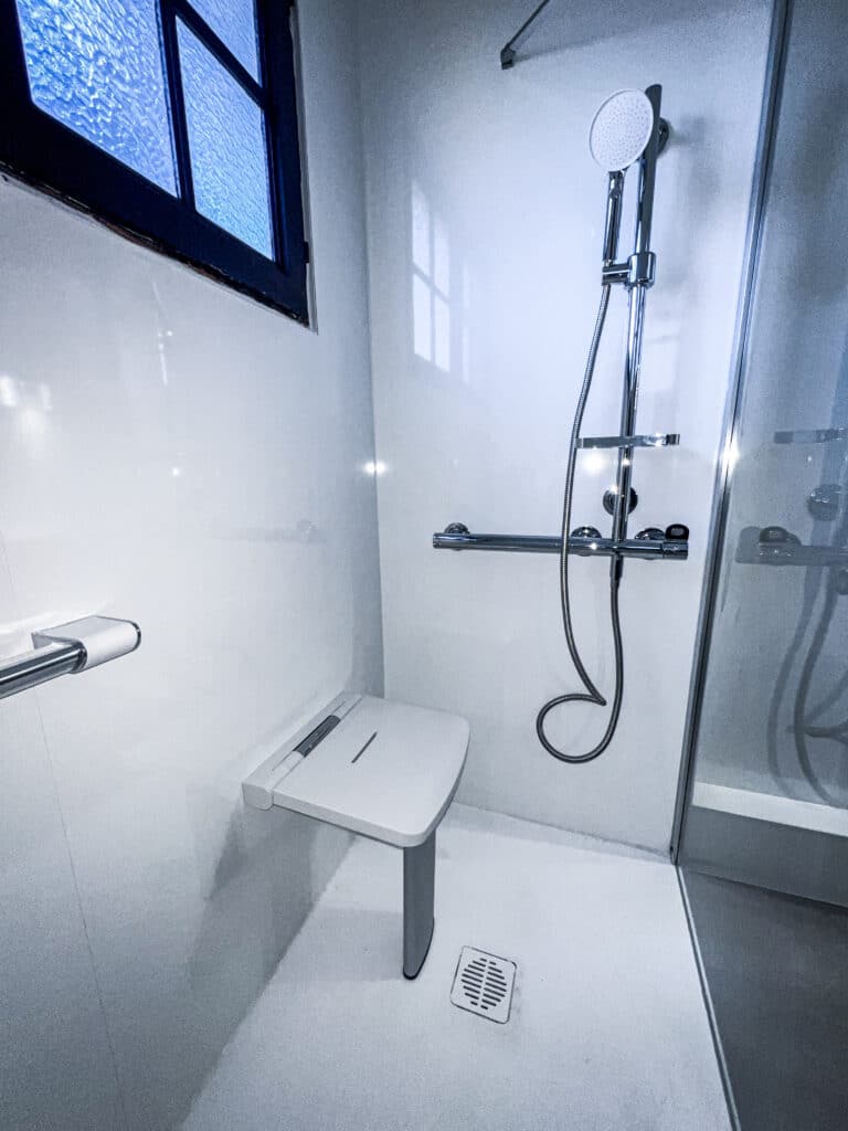 Douche PMR Moderne : Confort et Accessibilité Optimale Douche moderne et accessible avec siège rabattable mural, barre d'appui chromée, pommeau réglable et fenêtre à verre bleu texturé. Espace épuré.