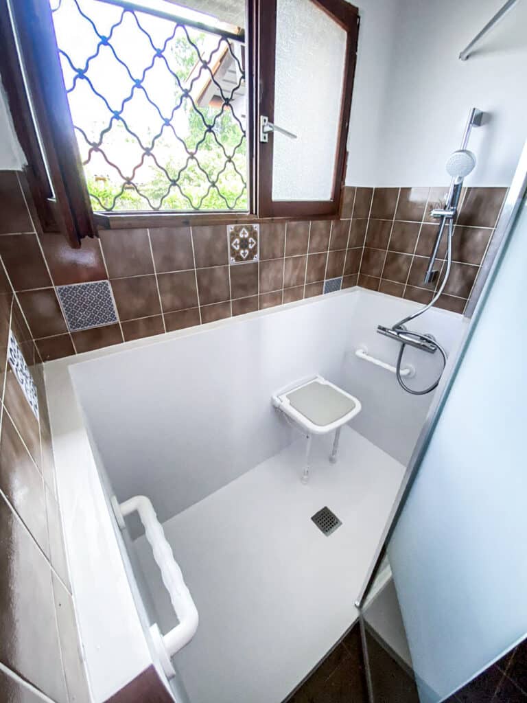 Salle de bain PMR moderne avec douche sécurisée, siège rabattable, barre d'appui, carrelage mural marron et fenêtre latérale.