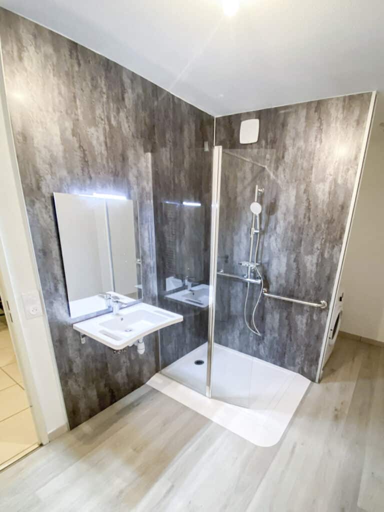 Salle de bain moderne et PMR avec murs gris texturés, lavabo suspendu, miroir lumineux et douche à l'italienne avec barres d'appui.