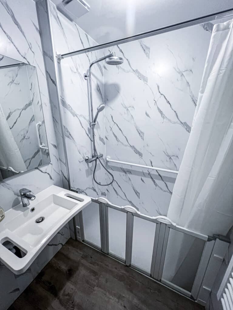 Salle de bain moderne blanche avec murs marbrés, lavabo, douche pluie, douchette, barre d'appui, rideau et écran pliant. Sol imitation bois foncé.