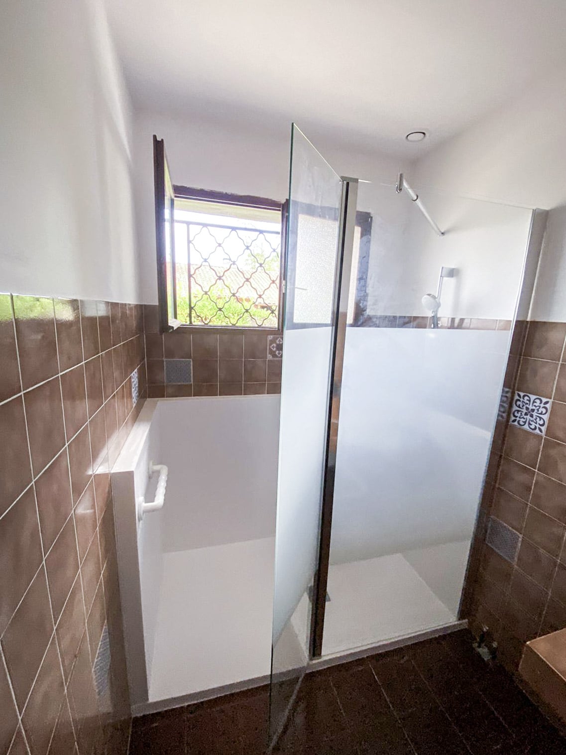 Salle de bain moderne: douche vitrée à l'italienne, baignoire-douche profonde avec barre d'appui. Murs carrelés bruns, fenêtre à grille.