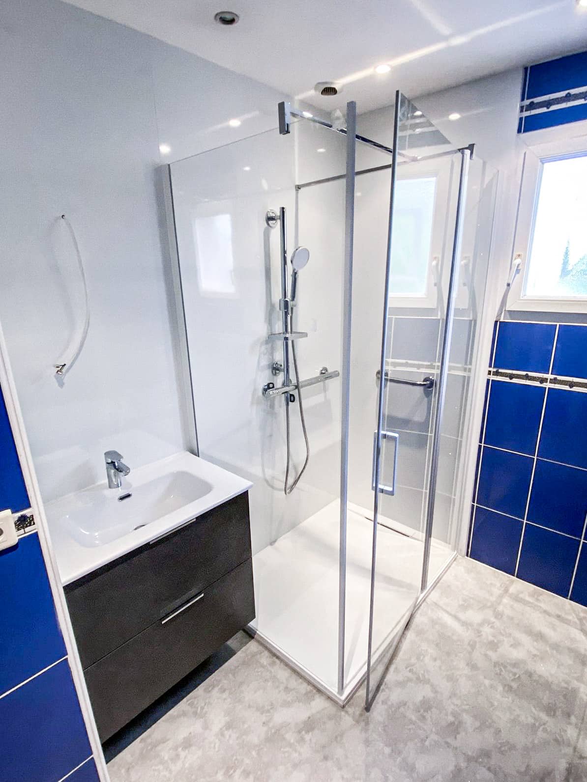 Salle de bain moderne: douche à l'italienne vitrée, meuble vasque foncé, mur carrelé bleu vif, sol béton ciré clair, et fenêtres lumineuses.