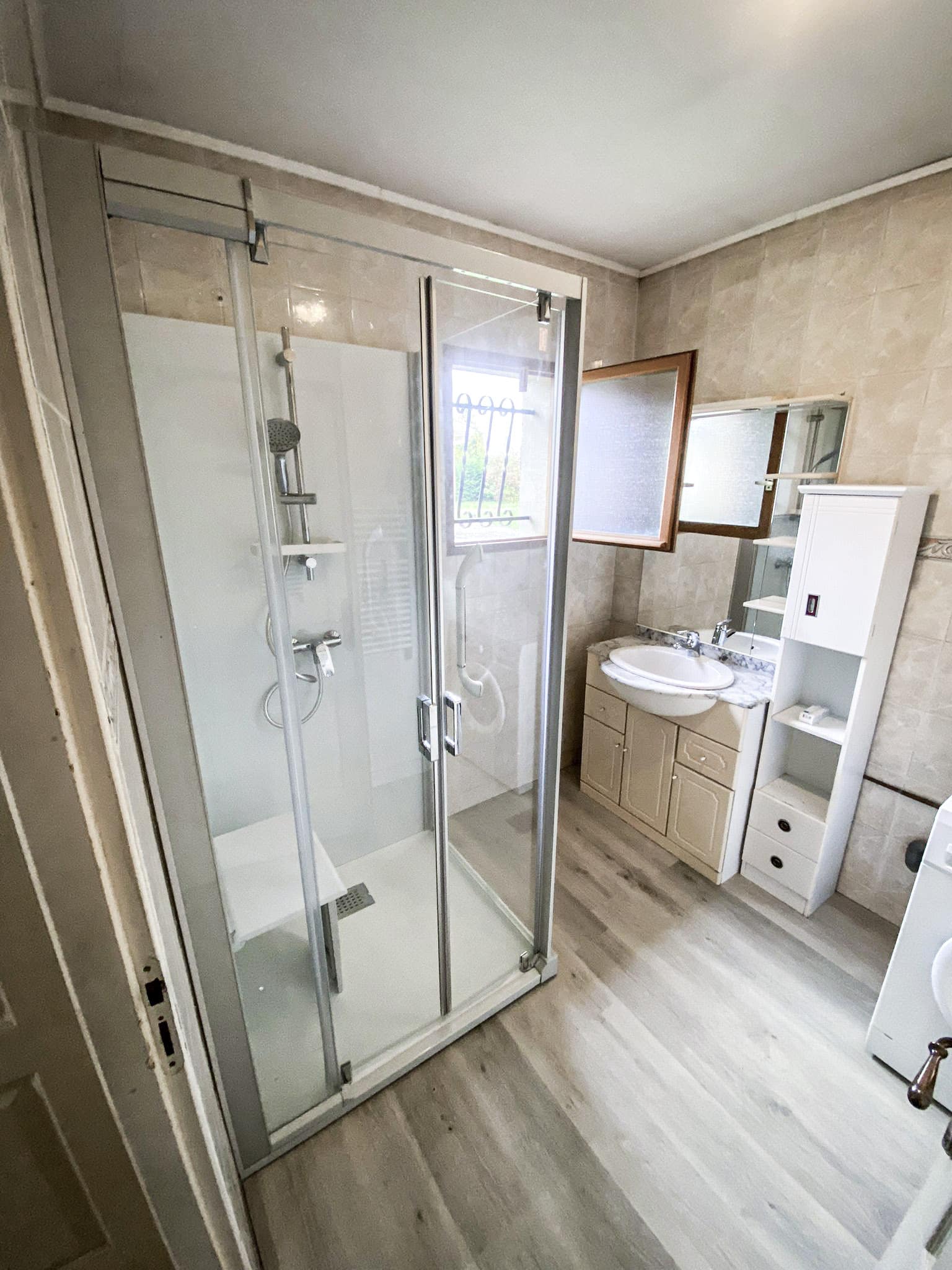 Salle de bain moderne: cabine de douche vitrée avec siège, meuble vasque, miroir, fenêtre, et colonne de rangement blanche. Sol clair.
