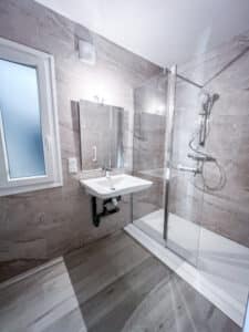 Salle de bain moderne avec carrelage mural effet marbre gris clair, sol aspect bois. Douche en verre, lavabo mural blanc, fenêtre givrée et miroir éclairé.