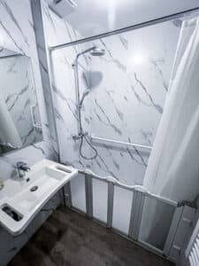 Salle de bain moderne: murs marbre blanc, lavabo, douche avec pommeau pluie, douchette, barres d'appui, rideau et accès PMR. Sol bois.