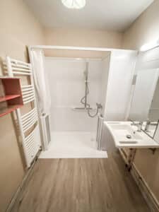 Salle de bain moderne beige. Douche à l'italienne accessible, barres d'appui, lavabo mural, miroir et sèche-serviettes blanc. Sol bois clair.
