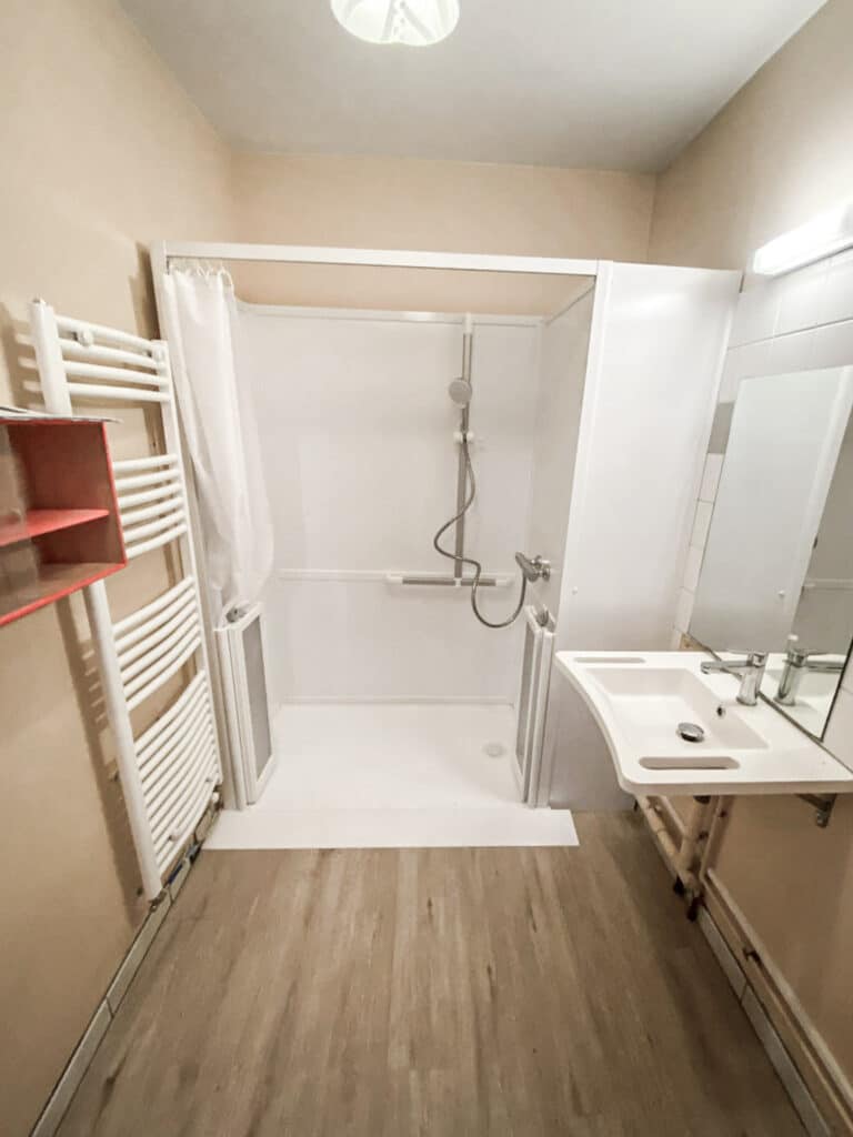 Salle de bain moderne beige. Douche à l'italienne accessible, barres d'appui, lavabo mural, miroir et sèche-serviettes blanc. Sol bois clair.