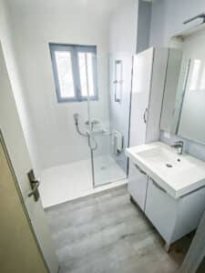 Salle de bain moderne blanche: douche à l'italienne, meuble vasque, miroir, grand placard et fenêtre à cadre bleu. Sol gris.