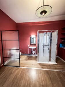 Studio moderne aux murs rouges avec salle de bain intégrée: douche vitrée, lavabo mural et WC derrière une partition. Plancher bois.