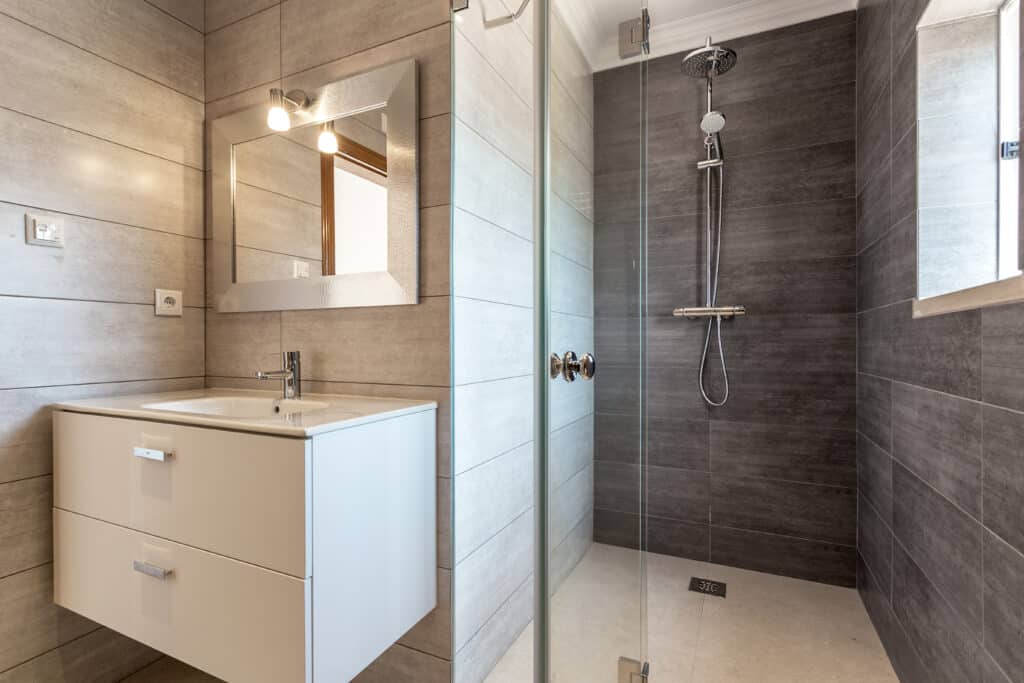 Salle de bain moderne carrelée beige et gris. Lavabo suspendu blanc, miroir argenté, douche vitrée avec pommeau pluie chromé et mélangeur.