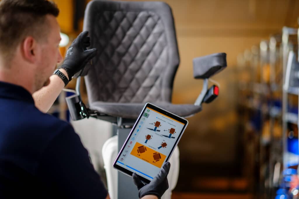 Contrôle numérique de fauteuil via tablette | Maintenance industrielle Un homme en gants noirs utilise une tablette pour contrôler un fauteuil gris matelassé. L'écran affiche 'Afstandsbediening' et les commandes 'Traction', 'Spindle', 'Swivel Motor'.