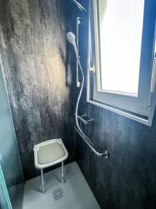 Douche accessible moderne avec murs sombres texturés. Comprend pommeau, barre d'appui chromée, siège rabattable blanc, et fenêtre latérale lumineuse.